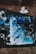 An abstract painting featuring swirling patterns of blue and black, accented with white splatters. The artwork appears to be on a canvas laid out on a dark surface, with paint cups and paper towels scattered nearby, suggesting a creative and spontaneous painting session.