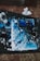 An abstract painting featuring swirling patterns of blue and black, accented with white splatters. The artwork appears to be on a canvas laid out on a dark surface, with paint cups and paper towels scattered nearby, suggesting a creative and spontaneous painting session.