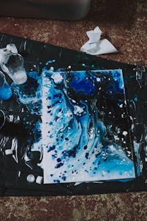 An abstract painting featuring swirling patterns of blue and black, accented with white splatters. The artwork appears to be on a canvas laid out on a dark surface, with paint cups and paper towels scattered nearby, suggesting a creative and spontaneous painting session.