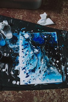 An abstract painting featuring swirling patterns of blue and black, accented with white splatters. The artwork appears to be on a canvas laid out on a dark surface, with paint cups and paper towels scattered nearby, suggesting a creative and spontaneous painting session.