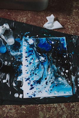 An abstract painting featuring swirling patterns of blue and black, accented with white splatters. The artwork appears to be on a canvas laid out on a dark surface, with paint cups and paper towels scattered nearby, suggesting a creative and spontaneous painting session.