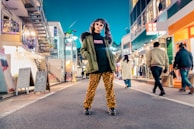 A person stands confidently in the middle of a vibrant city street lined with shops. They are wearing an olive green jacket with a fur collar, leopard print pants, black platform shoes, and sunglasses. Various pedestrians are walking by, and the area is brightly lit with colorful lights and signs from the shops. The atmosphere appears lively and stylish.