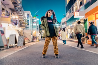 A person stands confidently in the middle of a vibrant city street lined with shops. They are wearing an olive green jacket with a fur collar, leopard print pants, black platform shoes, and sunglasses. Various pedestrians are walking by, and the area is brightly lit with colorful lights and signs from the shops. The atmosphere appears lively and stylish.