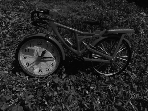 A small bicycle model is propped against a large clock placed on grass. The bicycle has intricate details, including visible handlebars, a seat, and spoked wheels. The clock displays the time with clear numbers and hands. The scene is monochrome, giving it a timeless, surreal quality.