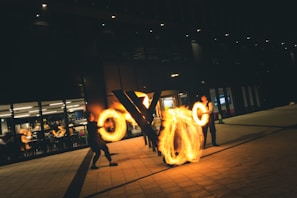 Performers are engaging in a fire juggling act on a dimly lit street, creating captivating circles and patterns with glowing fire. In the background, a modern building with large glass windows reveals people inside, possibly watching the performance from a café or restaurant.