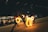 Performers are engaging in a fire juggling act on a dimly lit street, creating captivating circles and patterns with glowing fire. In the background, a modern building with large glass windows reveals people inside, possibly watching the performance from a café or restaurant.