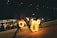 Performers are engaging in a fire juggling act on a dimly lit street, creating captivating circles and patterns with glowing fire. In the background, a modern building with large glass windows reveals people inside, possibly watching the performance from a café or restaurant.