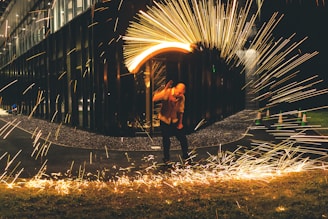 A person is surrounded by a dramatic display of bright, fiery sparks against a dark background. The path and building behind form a contrasting backdrop, while the motion of the sparks creates a dynamic and energetic effect.