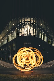 A modern building with illuminated windows and a large 'S4C' sign reflects a circular pattern of bright orange light trails created by a fire performer. The performance takes place on a pathway surrounded by greenery and small pebbles, adding an artistic dynamic to the urban setting.