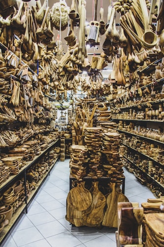 A store filled with numerous wooden items displayed on shelves and hanging from the ceiling. The assortment includes spoons, bowls, cutting boards, and various kitchen utensils made from wood. The arrangement creates a dense and rustic atmosphere.