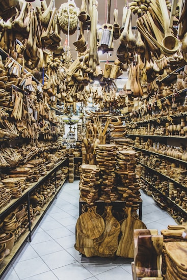 A store filled with numerous wooden items displayed on shelves and hanging from the ceiling. The assortment includes spoons, bowls, cutting boards, and various kitchen utensils made from wood. The arrangement creates a dense and rustic atmosphere.