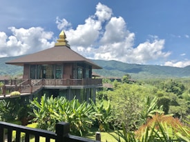 A wooden building with large windows is situated amidst lush greenery, with a backdrop of rolling mountains and a bright blue sky dotted with fluffy clouds. The architecture features a tiered roof with an ornamental spire. The surrounding landscape is rich with tall plants and trees, giving a sense of tranquility and natural beauty.