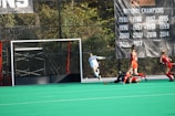 A field hockey match is in progress with several players actively moving near the goal. One player in a white uniform appears to be making a goal attempt, while three players in red have a defensive stance. A large banner listing past national championship years hangs in the background.