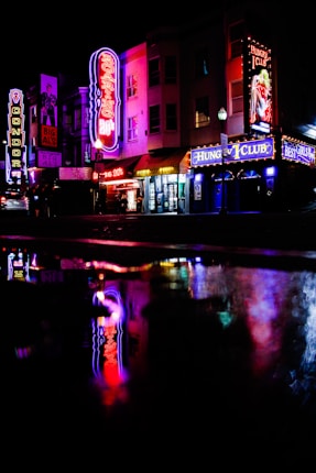 Neon signs illuminate a dark street, reflecting colorful lights onto the wet pavement. The scene captures a lively nightlife area with signs like 'Roaring 20's', 'Hungry Club', and 'Big Al's'.