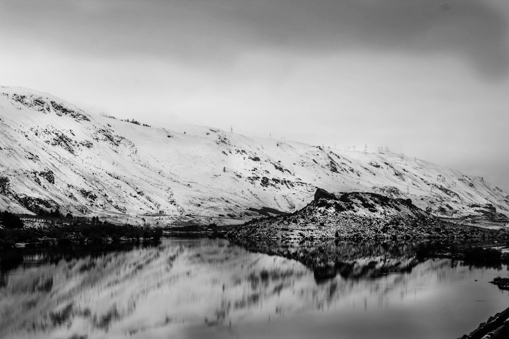 grayscale nature photography of body of water beside snow capped mountain under cloudy sky