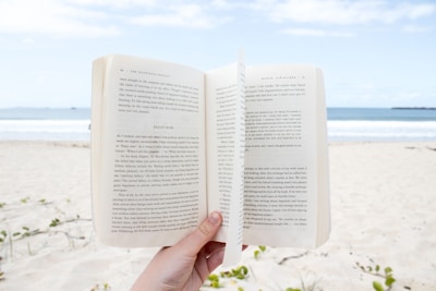 A person is holding an open book on a sandy beach with the ocean and a partly cloudy sky in the background. The scene captures a sense of relaxation and leisure by the seaside.