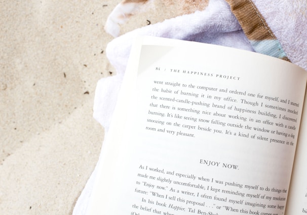 A book lies open on a sandy surface, with its pages exposed. The book's title, 'The Happiness Project,' is visible at the top of one page. A white towel is partially visible beneath the book, and the sand around it is light and textured with small grains.