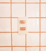 A switch panel is mounted on a tiled wall with white tiles and orange grout. The panel contains two switches labeled with hashtags: #selfie and #mood.