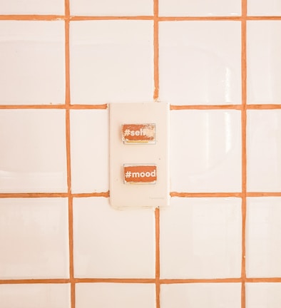 A switch panel is mounted on a tiled wall with white tiles and orange grout. The panel contains two switches labeled with hashtags: #selfie and #mood.