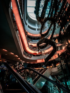 A modern multi-story shopping mall interior with escalators and lush greenery. Soft, warm lighting creates a welcoming atmosphere as decorative plants hang from the ceiling. Multiple levels are visible, showcasing a sleek and contemporary architectural design.
