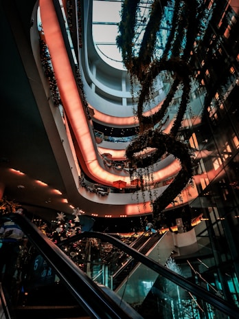 A modern multi-story shopping mall interior with escalators and lush greenery. Soft, warm lighting creates a welcoming atmosphere as decorative plants hang from the ceiling. Multiple levels are visible, showcasing a sleek and contemporary architectural design.
