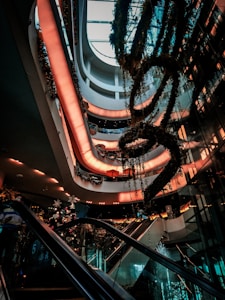 A modern multi-story shopping mall interior with escalators and lush greenery. Soft, warm lighting creates a welcoming atmosphere as decorative plants hang from the ceiling. Multiple levels are visible, showcasing a sleek and contemporary architectural design.
