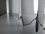 A close-up view of a row of vertical blinds with a bead chain. The blinds are positioned at a window with light filtering through, creating a soft, diffused light effect. The surface appears to be clean and the blinds have a smooth texture.