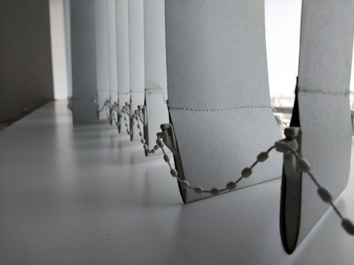 A close-up view of a row of vertical blinds with a bead chain. The blinds are positioned at a window with light filtering through, creating a soft, diffused light effect. The surface appears to be clean and the blinds have a smooth texture.