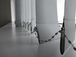 A close-up view of a row of vertical blinds with a bead chain. The blinds are positioned at a window with light filtering through, creating a soft, diffused light effect. The surface appears to be clean and the blinds have a smooth texture.