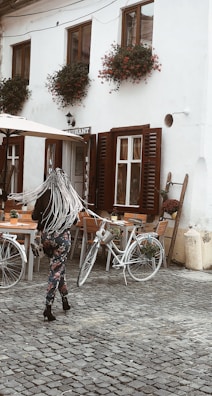 A person with long, braided hair is walking on a cobblestone pathway in front of a quaint café. The café has white walls, wooden shutters on the windows, and vibrant flower pots hanging from the windowsills. Two bicycles are parked near wooden tables and chairs outside the café.
