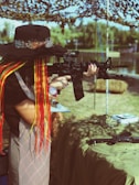 A person with long, colorful braided hair and a wide-brimmed hat aims a rifle in an outdoor setting. The background includes a camouflage net canopy, trees, and water, creating a serene yet focused environment.