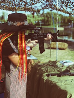 A person with long, colorful braided hair and a wide-brimmed hat aims a rifle in an outdoor setting. The background includes a camouflage net canopy, trees, and water, creating a serene yet focused environment.