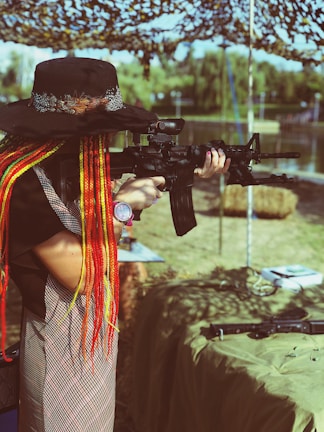 A person with long, colorful braided hair and a wide-brimmed hat aims a rifle in an outdoor setting. The background includes a camouflage net canopy, trees, and water, creating a serene yet focused environment.
