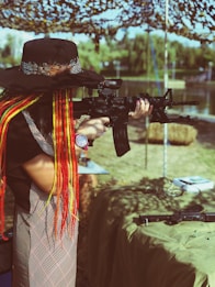 A person with long, colorful braided hair and a wide-brimmed hat aims a rifle in an outdoor setting. The background includes a camouflage net canopy, trees, and water, creating a serene yet focused environment.