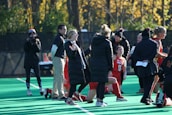 A group of people on a sports field, including several athletes in red uniforms and coaches wearing black coats. Some individuals are kneeling, while others are standing, engaged in animated conversation. A photographer or videographer is capturing the scene beside them. The background features a fence and autumn trees with yellow leaves.