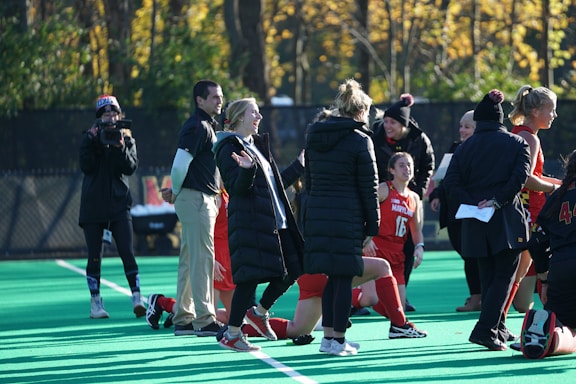 A group of people on a sports field, including several athletes in red uniforms and coaches wearing black coats. Some individuals are kneeling, while others are standing, engaged in animated conversation. A photographer or videographer is capturing the scene beside them. The background features a fence and autumn trees with yellow leaves.