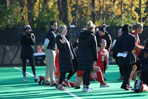 A group of people on a sports field, including several athletes in red uniforms and coaches wearing black coats. Some individuals are kneeling, while others are standing, engaged in animated conversation. A photographer or videographer is capturing the scene beside them. The background features a fence and autumn trees with yellow leaves.
