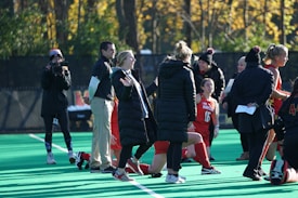 A group of people on a sports field, including several athletes in red uniforms and coaches wearing black coats. Some individuals are kneeling, while others are standing, engaged in animated conversation. A photographer or videographer is capturing the scene beside them. The background features a fence and autumn trees with yellow leaves.