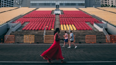 An open-air stadium with red and yellow seats arranged in rows on concrete steps. Three people are walking in front of the seating area, one in a vibrant red outfit, and two in casual sportswear. The background includes a modern building, and orange barriers are set up along the walkway.