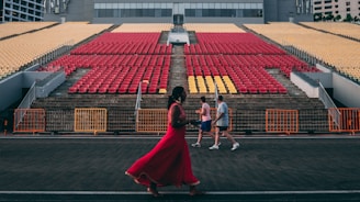 An open-air stadium with red and yellow seats arranged in rows on concrete steps. Three people are walking in front of the seating area, one in a vibrant red outfit, and two in casual sportswear. The background includes a modern building, and orange barriers are set up along the walkway.