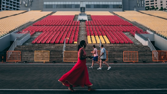 An open-air stadium with red and yellow seats arranged in rows on concrete steps. Three people are walking in front of the seating area, one in a vibrant red outfit, and two in casual sportswear. The background includes a modern building, and orange barriers are set up along the walkway.