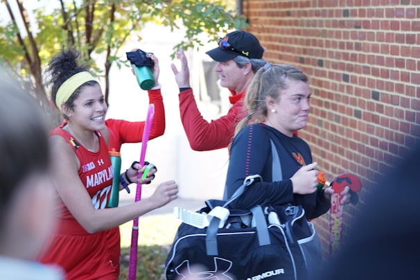 A group of athletes in sports attire appears to be in a joyful mood. One person is holding a field hockey stick and wearing a headband, while another carries a sports bag. They are gathered near a brick wall with trees in the background, sharing a light moment, likely after a sports event.