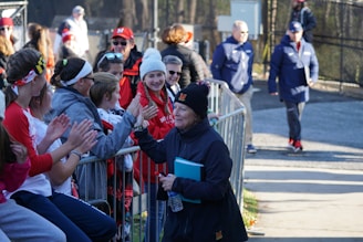 A group of people dressed in athletic and casual apparel stand near a metallic barricade, engaging in conversation and smiles. One person is extending a hand, possibly for a handshake or high-five. The setting appears to be outdoors near a sports field or event, with several individuals wearing caps or beanies to keep warm.