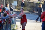 A female athlete dressed in a red sports uniform smiles while high-fiving a group of enthusiastic children and adults behind a metal barricade. The scene takes place outdoors with people wearing winter clothing, indicating a cold day.