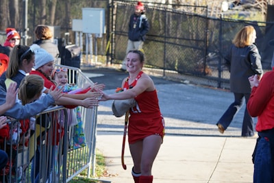 A female athlete dressed in a red sports uniform smiles while high-fiving a group of enthusiastic children and adults behind a metal barricade. The scene takes place outdoors with people wearing winter clothing, indicating a cold day.