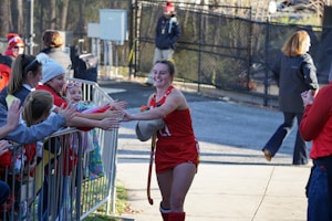 A female athlete dressed in a red sports uniform smiles while high-fiving a group of enthusiastic children and adults behind a metal barricade. The scene takes place outdoors with people wearing winter clothing, indicating a cold day.