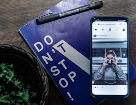 A smartphone showing a social media post featuring a person drinking from a cup. The phone rests on a blue book with the phrase 'DON'T STOP!' on it. A black pen is placed on the book, and part of a green plant is visible in the foreground.