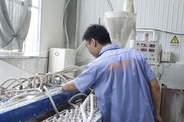 A person is working in an industrial setting, wearing a blue shirt with orange lettering on the back. There are multiple pipes and tubes connected to machinery. The room features industrial equipment like control panels with digital displays and a yellow caution sign. The walls are lined and there's a window draped with fabric.
