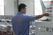 A person wearing a blue shirt operates a control panel in an industrial setting. The room features various machinery and control units with buttons and displays. Natural light enters through the windows, illuminating the workspace.