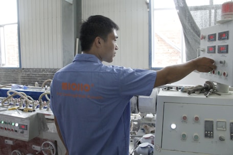 A person wearing a blue shirt operates a control panel in an industrial setting. The room features various machinery and control units with buttons and displays. Natural light enters through the windows, illuminating the workspace.